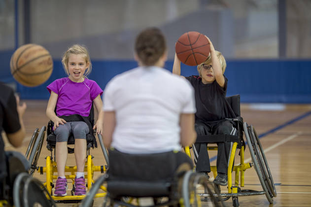Two disabled children are bouncing a basketball in the gym with their physical therapist.