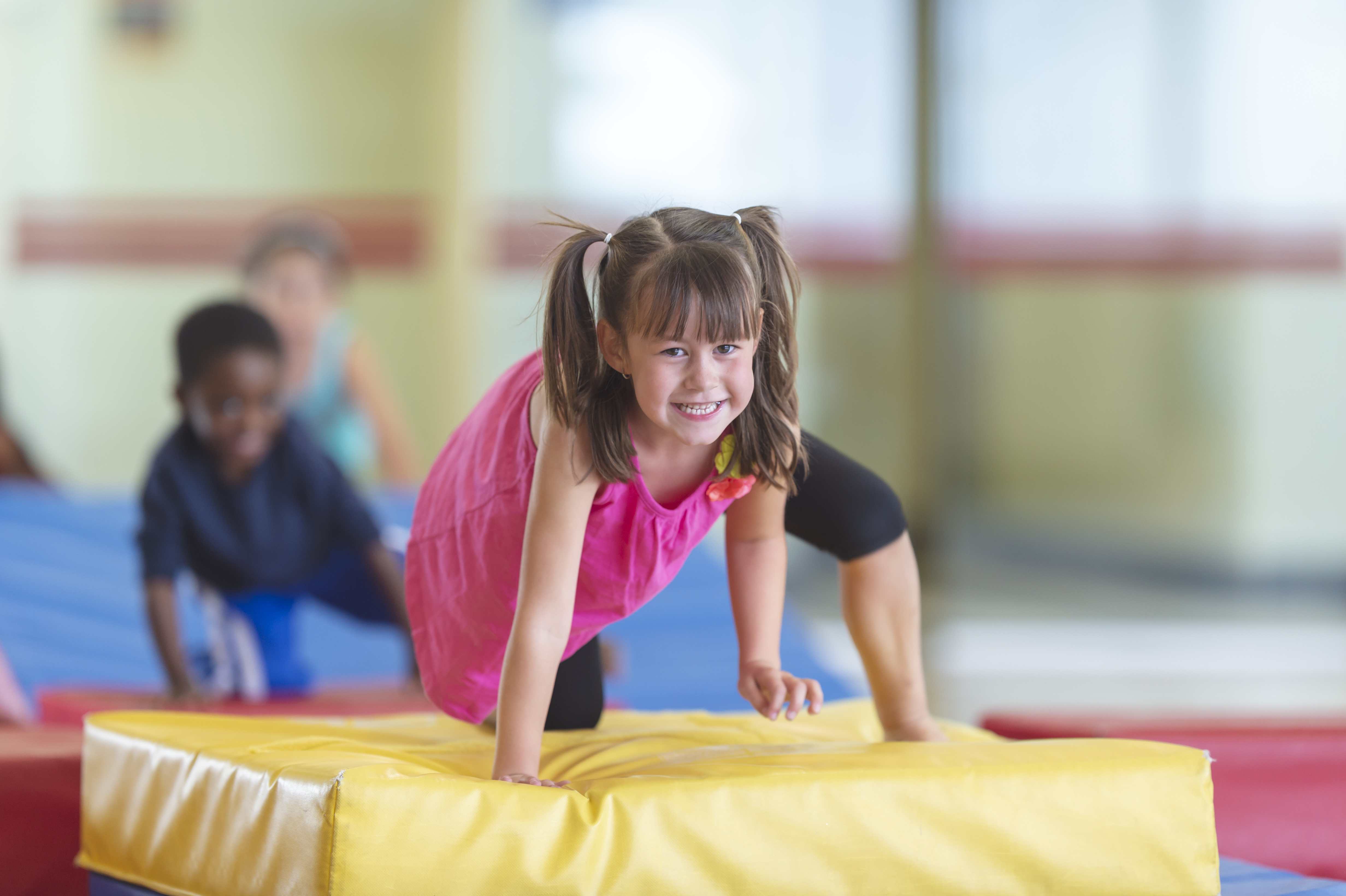 A young girl is smiling as she climbs a gymnastics landing pad. There are other children behind her. They are in a gymnasium.