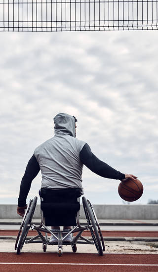Rug-aanzicht van basketter in rolstoel die dribbelt met bal outdoor, bewolkte lucht 