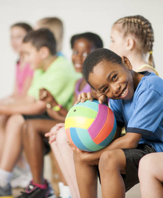 A multi-ethnic group of elementary age children are sitting in gym class and are going to play a game of volleyball. One boy is smiling while looking at the camera.