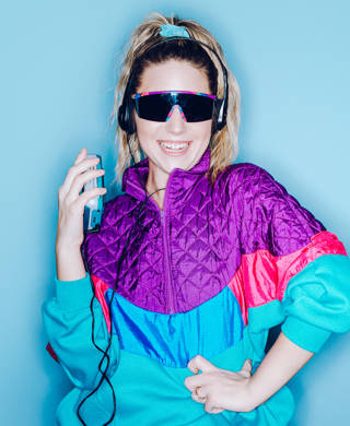 A woman wearing clothing styled after the 1980's and 1990's listens to music on her personal cassette tape player in front of a large bright blue background. Shot with a ring flash.